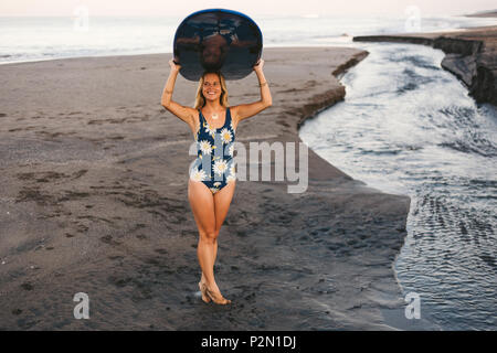 smiling sportswoman in swimming suit carrying blue surfing board on head on coastline Stock Photo