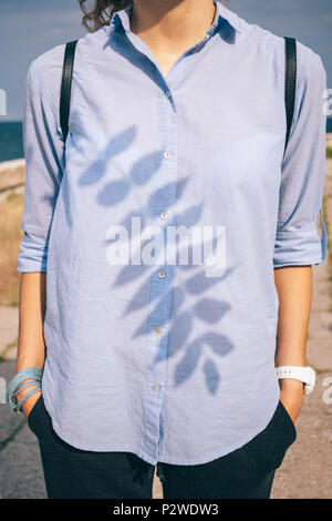Young woman standing by the sea, holding hands in her pockets, close-up. Shadow of a branch with leaves falls on a female's shirt on a sunny day. Stock Photo