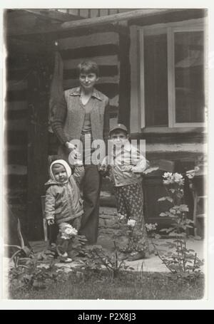 THE CZECHOSLOVAK  SOCIALIST REPUBLIC - CIRCA 1980s: Vintage photo shows an adolescent  girl with two children. They pose in front of log cabin. Retro black & white  photography. Stock Photo