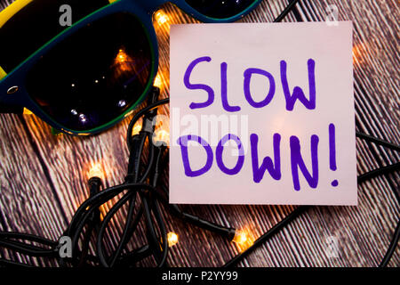 Slow Down handwritten on light pink note paper with blue color. Two sunglasses in different colors with wooden background and some lights are spread.  Stock Photo