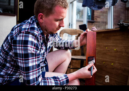 Painting old wooden furniture. Housekeeper paints wooden boards with painting brush. Home improvements and renovations concept. Young man holds painti Stock Photo