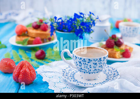 Small cupcakes with berries, strawberries and cherries. Delicious sweet breakfast and fragrant coffee, on a blue wooden background and flowers. Copy space. Stock Photo