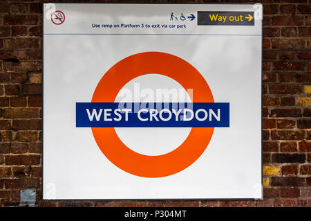 London overground tube style railway sign for West Croydon station.  White text on blue stripe, over red circle, on a white background, positioned against a brick wall. Stock Photo