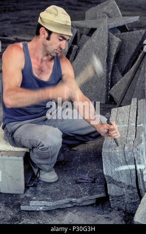 A professional stonecutter hammers a chisel along a straight line in a block of granite to separate a slab of the stone at a quarry in Riveo, a town in the Maggia Valley of Ticino, the Italian region of Switzerland. To cut stone slabs, the stonemason uses chisels with wide flat blades to tap along the cut line to produce a groove, then the chisels in that groove are hit harder to crack open the block of granite. This traditional method of stonecutting has been replaced by electric saws in many quarries. Stock Photo