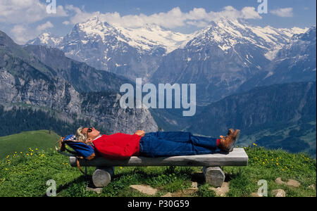 A female hiker rests on a wooden bench conveniently located along a trail that offers panoramic views of Jungfrau and other mountain summits in the Bernese Oberland region of Switzerland. The walking route to Schynige Platte plateau winds among alpine wildflowers that appear in summer after the snow melts in the higher altitudes of the Swiss Alps. Stock Photo