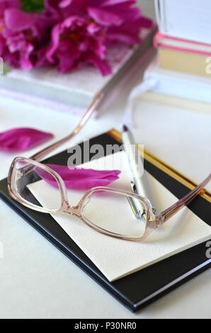 Bold violet pink peonies with blush pink glasses on a desk. Elegant feminine styled desktop workspace. Concept of working from home office, studying. Stock Photo