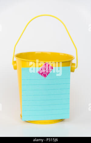 Aqua colored lined paper list taped to front of bright yellow miniature bucket with solid white background to illustrate bucket list Stock Photo