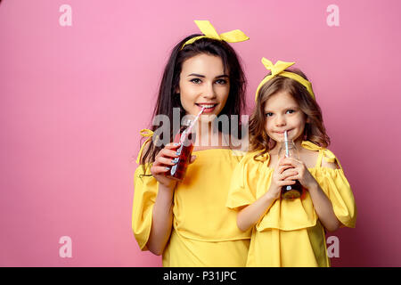 Crop view of attractive young woman with charming little girl in same yellow dresses holding red beverages smiling and looking at camera in studio on pink background. Stock Photo