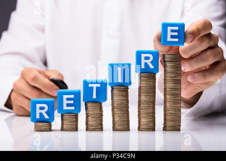 Person's Hand Making Retire Word By Placing Blue Cubic Blocks On Stacked Coins Over Desk Stock Photo