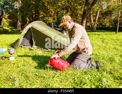 elderly tourist foldjng a tent in the forest on sunny summer day Stock Photo