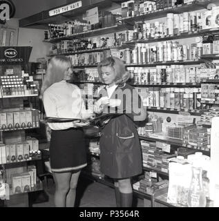 1970, two young ladies in a chemist's, one with long hair and wearing a short skirt, showing the other, in a rainmac, the range of cosmetic and hair products available, England, UK. Stock Photo