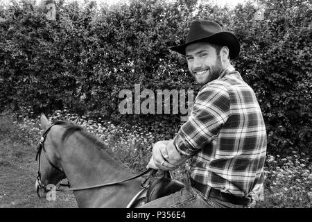 Good looking, hunky cowboy rides horse with boots, chequered shirt and hat Stock Photo