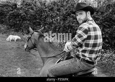 Good looking, hunky cowboy rides horse with boots, chequered shirt and hat Stock Photo