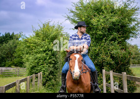 Good looking, hunky cowboy rides horse with boots, chequered shirt and hat Stock Photo