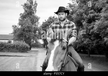 Good looking, hunky cowboy rides horse with boots, chequered shirt and hat Stock Photo