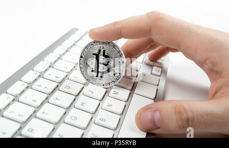 hand holds a silver coin of bitcoin above the keyboard Stock Photo
