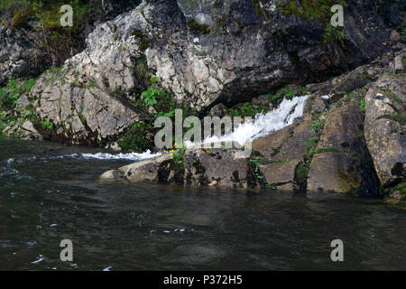 The spring follows a waterfall from the rock and flows into the river on a precipitous river bank Stock Photo