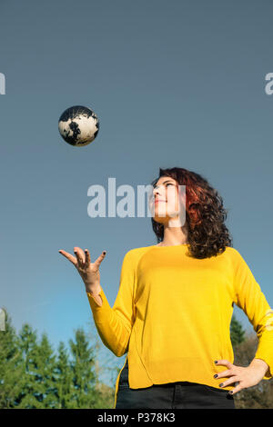 The whole world in her hand: Young woman in stylish yellow pullover catching earth in her hand in front of sky and trees. Save the earth concept. Stock Photo