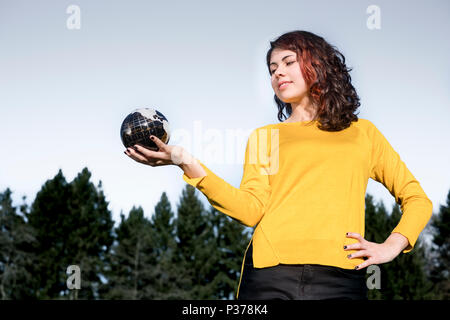 The whole world in her hand: Young woman in stylish yellow pullover holding earth in her hand in front of sky and trees. Save the earth concept. Stock Photo