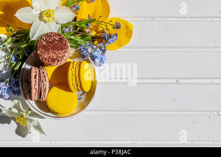 Colorful macaroons on plate on white wooden table. Sweet macarons and flowers. Composition with tender flowers blossoms and french dessert. Top view with copy space Stock Photo
