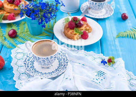 Small cupcakes with berries, strawberries and cherries. Delicious sweet breakfast and fragrant coffee, on a blue wooden background and flowers. Copy space. Stock Photo