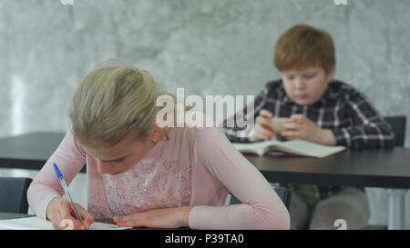 A young girl in a classroom concentrating on her test, while her classmate playing games on smartphone Stock Photo