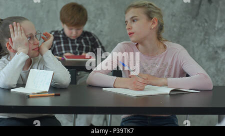 Bored girl sitting at desk in school classroom, discussing something with classmate Stock Photo
