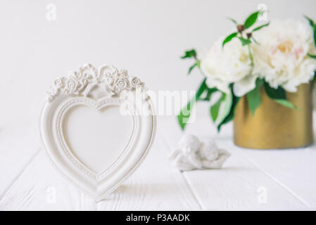 Heart shaped photoframe with plaster flowers, statuette of two antique little lovely angels on the white wooden table with bouquet of white peonies. L Stock Photo