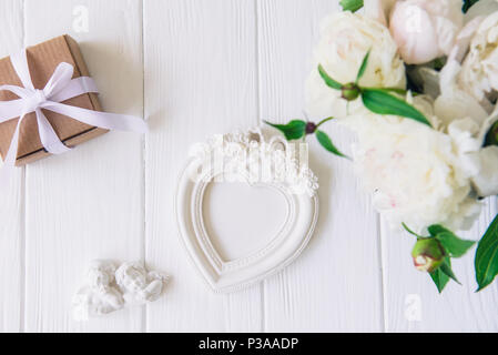 Top view vintage heart shaped photo frame, statuette of two antique lovely angels, giftbox and white peonies bouquet on the wooden table. Love backgro Stock Photo