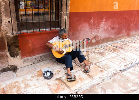 A crippled homeless busker playing guitar and harmonica, collecting money in a hat, in San Miguel de Allende Stock Photo