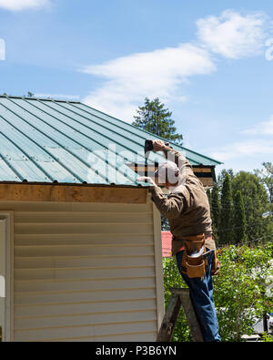 Construction worker on a ladder repairing damage to a green metal roof in Speculator, NY USA Stock Photo