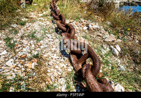 Old rusty ship anchor metal chain laying on the ground. Giant heavy steel chain leading from ground to the sea. Stock Photo