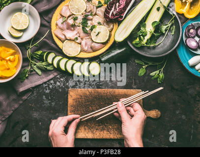 Chicken meat and vegetables skewers making. Female hands holding wooden skewer on kitchen table background with chicken pieces and vegetables, top vie Stock Photo