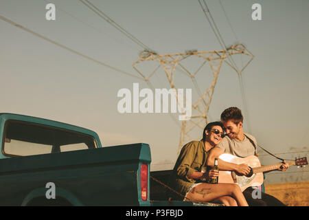 Couple sitting in the back of their pickup truck enjoying the road trip in country side. Man playing the guitar while the woman is holding a bottle of Stock Photo