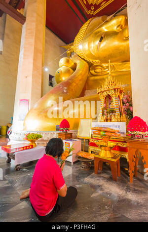 Phetchaburi, Thailand - March 2018: Red shirt woman worshiping reclining golden Buddha image with bunch of flowers and incense in Buddhist church in P Stock Photo