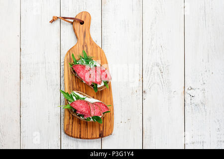 Tasty appetizer of thinly sliced spicy salami on bread with cheese and herbs on a cutting board with copy space Stock Photo