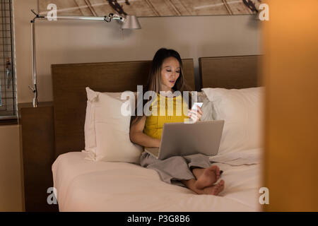 Businesswoman sitting on bed using her phone while working on laptop Stock Photo
