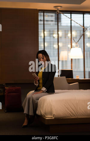 Businesswoman sitting on bed using her mobile phone Stock Photo