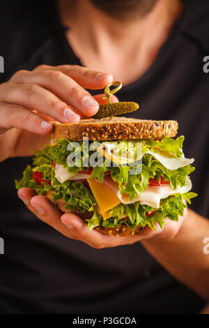 Big sandwich with bread, meat (porshutto), cheese and fresh vegetables in male hands, dark  background. Stock Photo