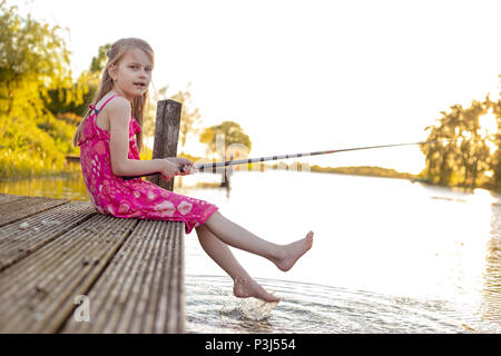 A 7 year old girl is sitting with her fishing rod on a jetty by a lake, waiting for a fish to bite. She looks into the camera and splashes with her fe Stock Photo