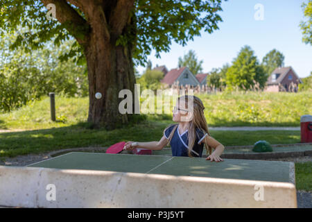 A 7 year old girl plays table tennis on an outside ping-pong table. She is focused on hitting the ball with her bat Stock Photo