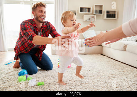 Toddler girl walking from dad to mum’s arms in sitting room Stock Photo