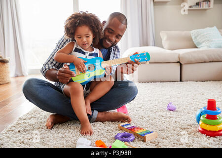 Toddler daughter sits on dad’s knee playing ukulele at home Stock Photo