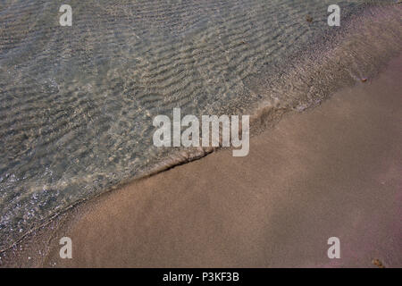 Small wave playing with pink toned sand of famous Elafonisi beach at Crete island, Greece. Wavy sand bottom can be clearly seen through clean water. Stock Photo