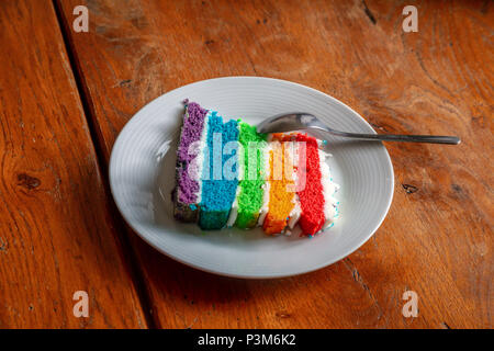 A slice of multicolored rainbow layer sponge cake on a white plate on a wooden table. Stock Photo