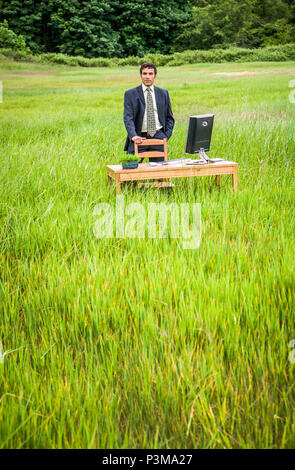A businessman standing behind a chair in a green grassy field at a desk with a computer on it. Green business concept. Stock Photo
