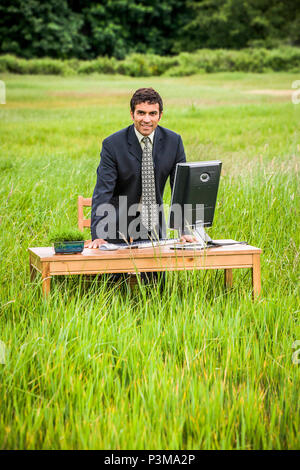 A businessman standing in a green field leaning on a desk with a computer on it. Green business concept. Stock Photo