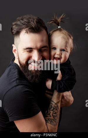 Handsome tattooed young man holding cute little baby on black background Stock Photo