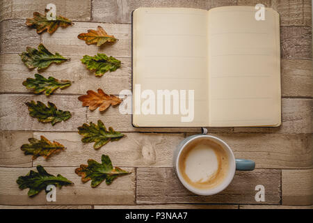 Top view of an open notebook and a cappuccino mug with green and orange leaves on a wooden board. Landscape format. Stock Photo