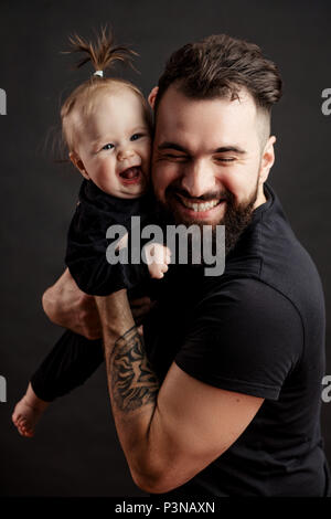 Handsome tattooed young man holding cute little baby on black background Stock Photo